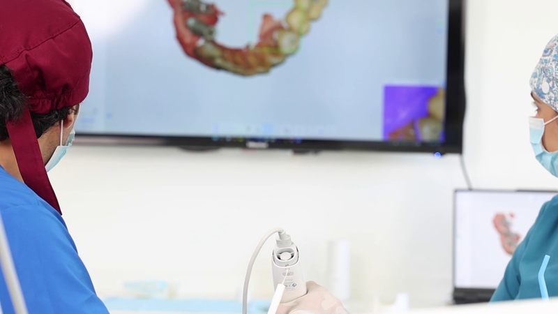 An intraoral scanner in use, with the scan building up on the screen behind
